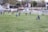 A football match is taking place on a grassy field with players wearing blue and black jerseys. One player lies on the ground while others are scattered around the field. There is an ambulance parked nearby, and flags are flying high. A referee in a bright uniform is present, and the background shows a few buildings and trees.