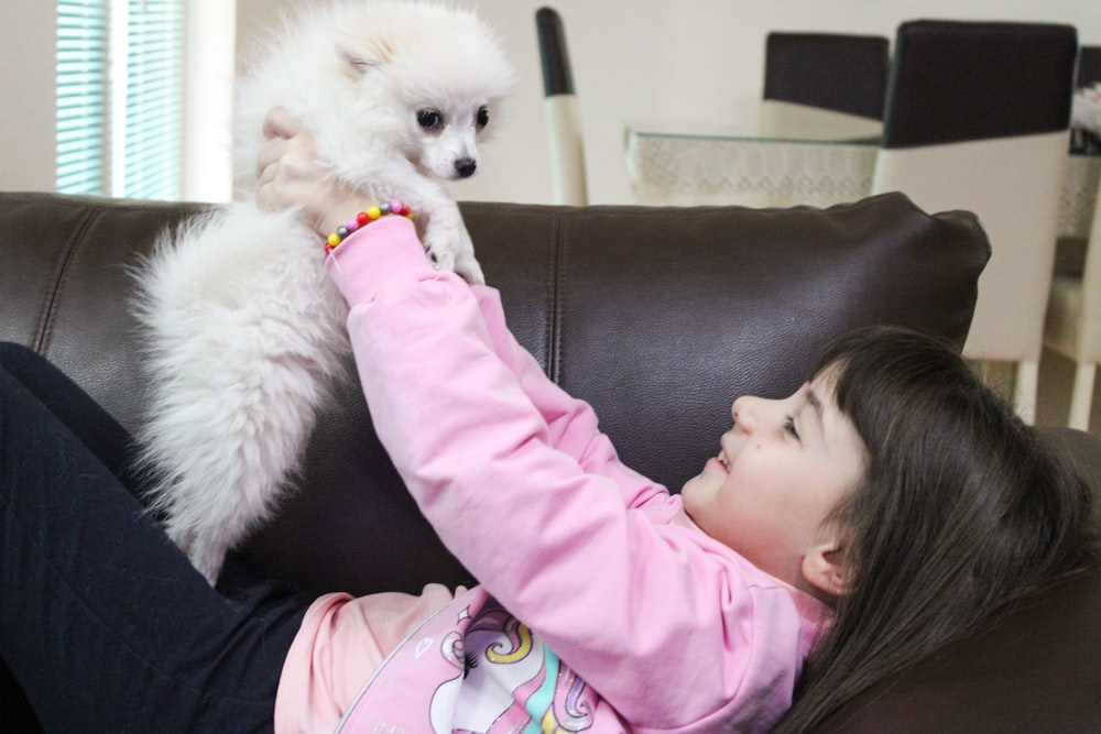 a little girl sitting on a couch petting a small white dog