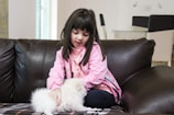 A young girl in a pink hoodie is sitting on a dark brown leather sofa petting a fluffy white kitten. The room has light-colored walls and some modern furniture in the background.