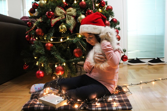 A child wearing a Santa hat sits on a checkered blanket near a decorated Christmas tree. The tree is adorned with red and gold ornaments, lights, and a bow. The child is holding a fluffy white object, likely a stuffed animal, and is surrounded by glowing string lights. A book lies open on the floor, and a pair of shoes is visible in the background.
