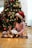 A child wearing a Santa hat is sitting on a checkered blanket in front of a decorated Christmas tree. The tree has red and gold ornaments, bows, and string lights. The child is holding an open book and is surrounded by lit Christmas lights on the floor.