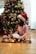 A child wearing a Santa hat is sitting on a checkered blanket in front of a decorated Christmas tree. The tree has red and gold ornaments, bows, and string lights. The child is holding an open book and is surrounded by lit Christmas lights on the floor.