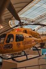 A yellow air ambulance helicopter parked indoors within a large exhibition hall. The helicopter has the label 'Tyrolean Air Ambulance' and various logos including 'ÖAMTC' and 'Christophorus'. The setting appears to be a museum or gallery, indicated by the architectural structure of the roof and surrounding displays.