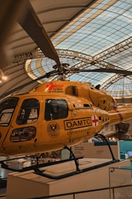 A yellow air ambulance helicopter parked indoors within a large exhibition hall. The helicopter has the label 'Tyrolean Air Ambulance' and various logos including 'ÖAMTC' and 'Christophorus'. The setting appears to be a museum or gallery, indicated by the architectural structure of the roof and surrounding displays.