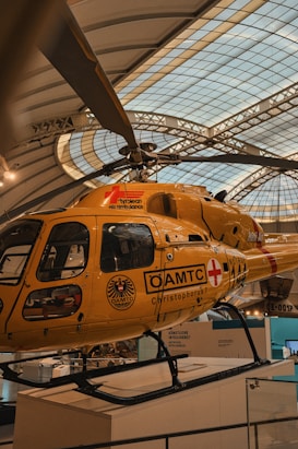 A yellow air ambulance helicopter parked indoors within a large exhibition hall. The helicopter has the label 'Tyrolean Air Ambulance' and various logos including 'ÖAMTC' and 'Christophorus'. The setting appears to be a museum or gallery, indicated by the architectural structure of the roof and surrounding displays.