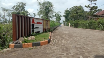 A paved pathway lined with greenery and shrubs leads into the distance. A prominent sign with large red and black letters stands to the left, surrounded by grass and small rocks. Trees and bushes are visible, contributing to a natural and serene setting.