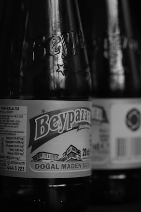 Close-up view of a black and white photograph showcasing bottles with labels. The prominent label reads 'Beypazarı,' indicating a brand of mineral water. Details such as nutritional information and a barcode are visible. The focus on the bottle creates a textured glass effect.
