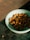 A light blue bowl contains triangular, brown pieces that appear to be pet food or snacks. The bowl is placed on a dark, woven mat, with a minimal piece scattered nearby.