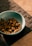 A blue bowl containing pieces of dry pet food is placed on a dark woven mat. The setting appears to be indoors with a tiled floor partially visible.