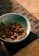 A blue bowl containing pieces of dry pet food is placed on a dark woven mat. The setting appears to be indoors with a tiled floor partially visible.