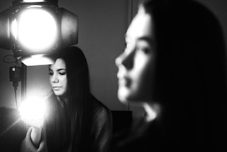 A black and white photograph of a woman sitting in front of a bright light source, holding a glowing object in her hand. The focus is on the woman, with a blurred silhouette of another person in the foreground.