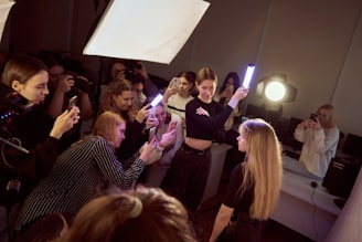 A group of people is gathered in an indoor setting, with several individuals focused on taking photographs using smartphones. One person is holding a light stick, illuminating a young woman with long hair, who is the apparent subject of the photoshoot. Bright lighting equipment is set up, enhancing the clarity of the scene.