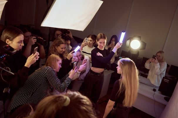 A group of people is gathered in an indoor setting, with several individuals focused on taking photographs using smartphones. One person is holding a light stick, illuminating a young woman with long hair, who is the apparent subject of the photoshoot. Bright lighting equipment is set up, enhancing the clarity of the scene.