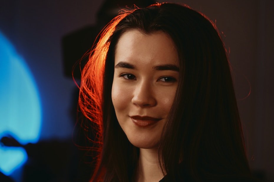 A close-up portrait of a woman with long dark hair, softly lit with warm tones. Her hair is illuminated with a vivid red highlight, creating a striking contrast against the dimly lit background with a blue glow.