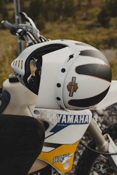 A white motorcycle helmet is resting on a Yamaha dirt bike, displaying logos and designs. The bike, situated in an outdoor setting, shows rugged terrain in the background, suggesting an off-road context.