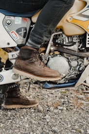 A person wearing dark jeans and brown leather boots sits on a Yamaha dirt bike. The bike has various stickers on its body, including branding and logos. The ground is covered with small rocks and gravel, adding an outdoor, rugged feel.