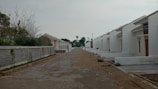A row of newly constructed white houses lined up along a paved street. The homes feature simple architectural designs with slanted roofs. On one side, there is a tall concrete wall with sparse vegetation. The ground appears to be under development with exposed earth and some construction debris.