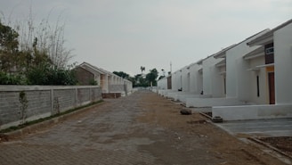 A row of newly constructed white houses lined up along a paved street. The homes feature simple architectural designs with slanted roofs. On one side, there is a tall concrete wall with sparse vegetation. The ground appears to be under development with exposed earth and some construction debris.
