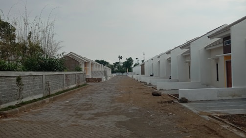 A row of newly constructed white houses lined up along a paved street. The homes feature simple architectural designs with slanted roofs. On one side, there is a tall concrete wall with sparse vegetation. The ground appears to be under development with exposed earth and some construction debris.