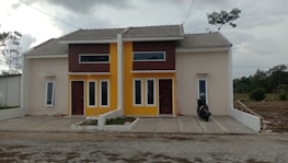 A pair of modern, minimalist houses side by side with a beige and brown exterior, featuring a shared roof and driveway. The building has large vertical windows and one of them is painted with a bright yellow accent. On the right side of the driveway, a parked motorcycle is visible. The surrounding area is somewhat overcast with scattered trees and greenery.