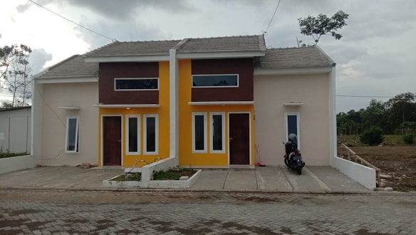 A pair of modern, minimalist houses side by side with a beige and brown exterior, featuring a shared roof and driveway. The building has large vertical windows and one of them is painted with a bright yellow accent. On the right side of the driveway, a parked motorcycle is visible. The surrounding area is somewhat overcast with scattered trees and greenery.