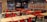 A fast-food restaurant interior with empty tables and chairs. The seating consists of a mix of red and black metal chairs and benches, with light wood tabletops. The wall features a large logo and a slogan in bold lettering. Several pendant lights hang from the ceiling, reflecting the establishment's modern design. The restaurant is well-lit, giving it a clean and inviting atmosphere.