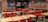 A fast-food restaurant interior with empty tables and chairs. The seating consists of a mix of red and black metal chairs and benches, with light wood tabletops. The wall features a large logo and a slogan in bold lettering. Several pendant lights hang from the ceiling, reflecting the establishment's modern design. The restaurant is well-lit, giving it a clean and inviting atmosphere.