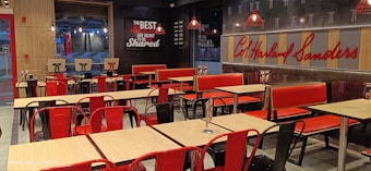 A fast-food restaurant interior with empty tables and chairs. The seating consists of a mix of red and black metal chairs and benches, with light wood tabletops. The wall features a large logo and a slogan in bold lettering. Several pendant lights hang from the ceiling, reflecting the establishment's modern design. The restaurant is well-lit, giving it a clean and inviting atmosphere.