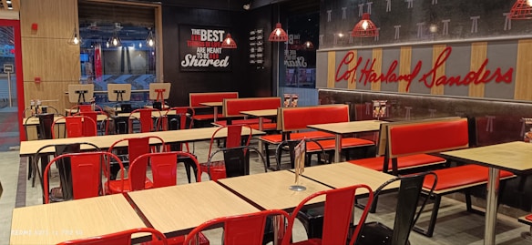 A fast-food restaurant interior with empty tables and chairs. The seating consists of a mix of red and black metal chairs and benches, with light wood tabletops. The wall features a large logo and a slogan in bold lettering. Several pendant lights hang from the ceiling, reflecting the establishment's modern design. The restaurant is well-lit, giving it a clean and inviting atmosphere.