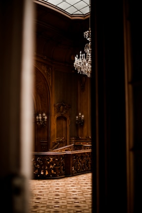 An ornate interior featuring intricate woodwork and a grand chandelier hanging from the ceiling. The space is elegantly lit, highlighting the detailed craftsmanship of the wooden staircase and decorative moldings.