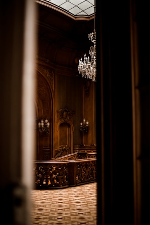 An ornate interior featuring intricate woodwork and a grand chandelier hanging from the ceiling. The space is elegantly lit, highlighting the detailed craftsmanship of the wooden staircase and decorative moldings.