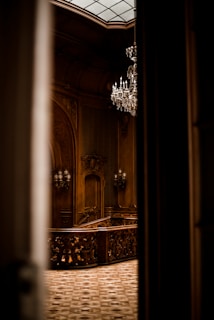 An ornate interior featuring intricate woodwork and a grand chandelier hanging from the ceiling. The space is elegantly lit, highlighting the detailed craftsmanship of the wooden staircase and decorative moldings.