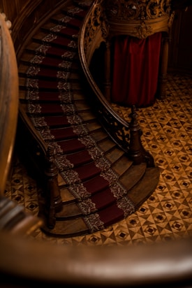 An ornate wooden staircase with a red patterned carpet runner. The staircase curves elegantly, with intricately carved banisters and an ornate wooden design. The floor features a geometric pattern and a rich, warm color palette. The lighting is dim, adding a sense of history and grandeur.