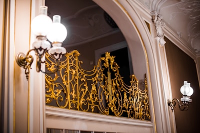 Ornate golden railing with intricate designs is mounted on a sophisticated interior wall. The surroundings include classic light fixtures with frosted globes that emit a warm glow. The decor exudes elegance and opulence, with subtle carvings and molding on the walls.