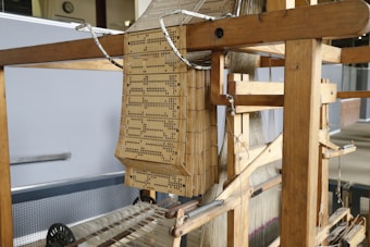 A traditional wooden loom featuring perforated punch cards used for weaving patterns. The punch cards are intricately arranged, showcasing their use in guiding the loom's process. The structure is made primarily of wood, with visible threads arranged neatly for weaving.