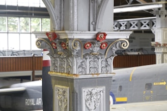 An ornate architectural column with intricate designs and decorative elements, featuring red and gold accents. In the background, part of a large aircraft with the text 'ROYAL AIR FORCE' is visible in a hangar or museum setting.