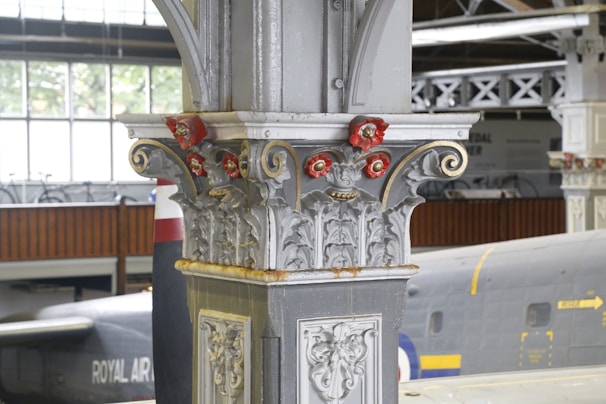An ornate architectural column with intricate designs and decorative elements, featuring red and gold accents. In the background, part of a large aircraft with the text 'ROYAL AIR FORCE' is visible in a hangar or museum setting.