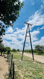 A rural landscape with a dirt road running alongside power lines supported by wooden poles. The road is bordered by patches of green vegetation and trees. A rustic wire fence is visible in the foreground. The sky is bright with scattered clouds.