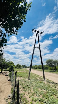A rural landscape with a dirt road running alongside power lines supported by wooden poles. The road is bordered by patches of green vegetation and trees. A rustic wire fence is visible in the foreground. The sky is bright with scattered clouds.