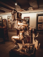 A metallic sculpture depicting two intertwined human figures giving an impression of movement and complexity, placed in an art gallery surrounded by framed artworks and ambient lighting.