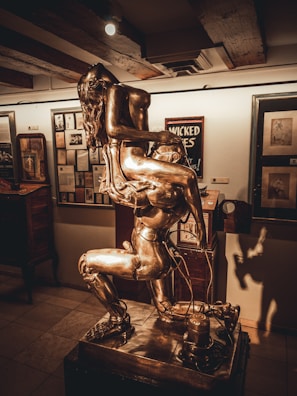 A metallic sculpture depicting two intertwined human figures giving an impression of movement and complexity, placed in an art gallery surrounded by framed artworks and ambient lighting.