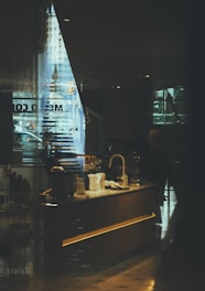 A dimly lit cafe interior with a modern design. The reflection of an urban street scene can be seen through large glass windows, creating a blend of indoor and outdoor elements. The counter features a sink and various items, possibly for serving drinks or food. The atmosphere suggests a cozy, inviting place for visitors.