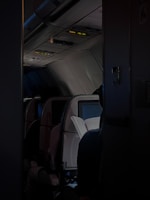 A dimly lit airplane cabin with a focus on a passenger reading a book in their seat. The overhead bins and ceiling controls are visible, and the seatback in-flight entertainment screens are turned off, adding to a calm and quiet atmosphere.