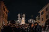 A crowd gathered in a bustling square during the night, with a large illuminated Christmas tree in the center. Historical buildings tower in the background, including a church with twin bell towers. The atmosphere is filled with warmth and festivity as people engage with each other, wrapped in winter clothing.