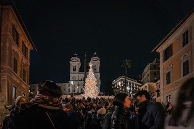 A crowd gathered in a bustling square during the night, with a large illuminated Christmas tree in the center. Historical buildings tower in the background, including a church with twin bell towers. The atmosphere is filled with warmth and festivity as people engage with each other, wrapped in winter clothing.