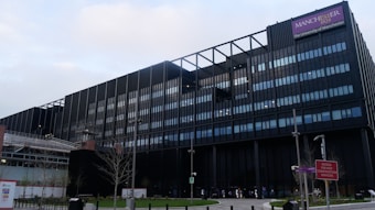 A large modern building with a predominantly black exterior and glass windows. Scaffolding is visible on one side, suggesting construction or renovation work. A sign reads 'Manchester' and 'The University of Manchester' in the upper right corner. There are leafless trees, a construction sign, benches, and street lamps in front of the structure.
