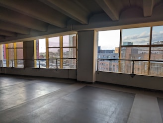 An empty parking garage with concrete flooring and ceiling beams. The space is partially enclosed, with large windows bordered by yellow frames. Outside, a cityscape is visible, featuring several buildings under a cloudy sky.