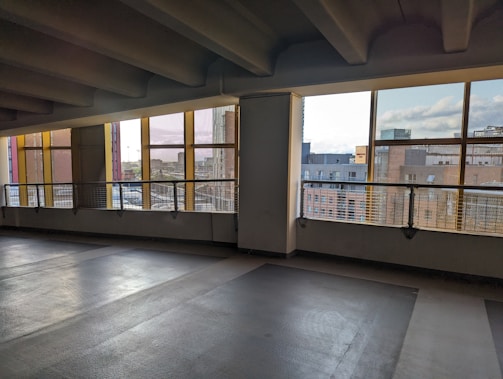 An empty parking garage with concrete flooring and ceiling beams. The space is partially enclosed, with large windows bordered by yellow frames. Outside, a cityscape is visible, featuring several buildings under a cloudy sky.