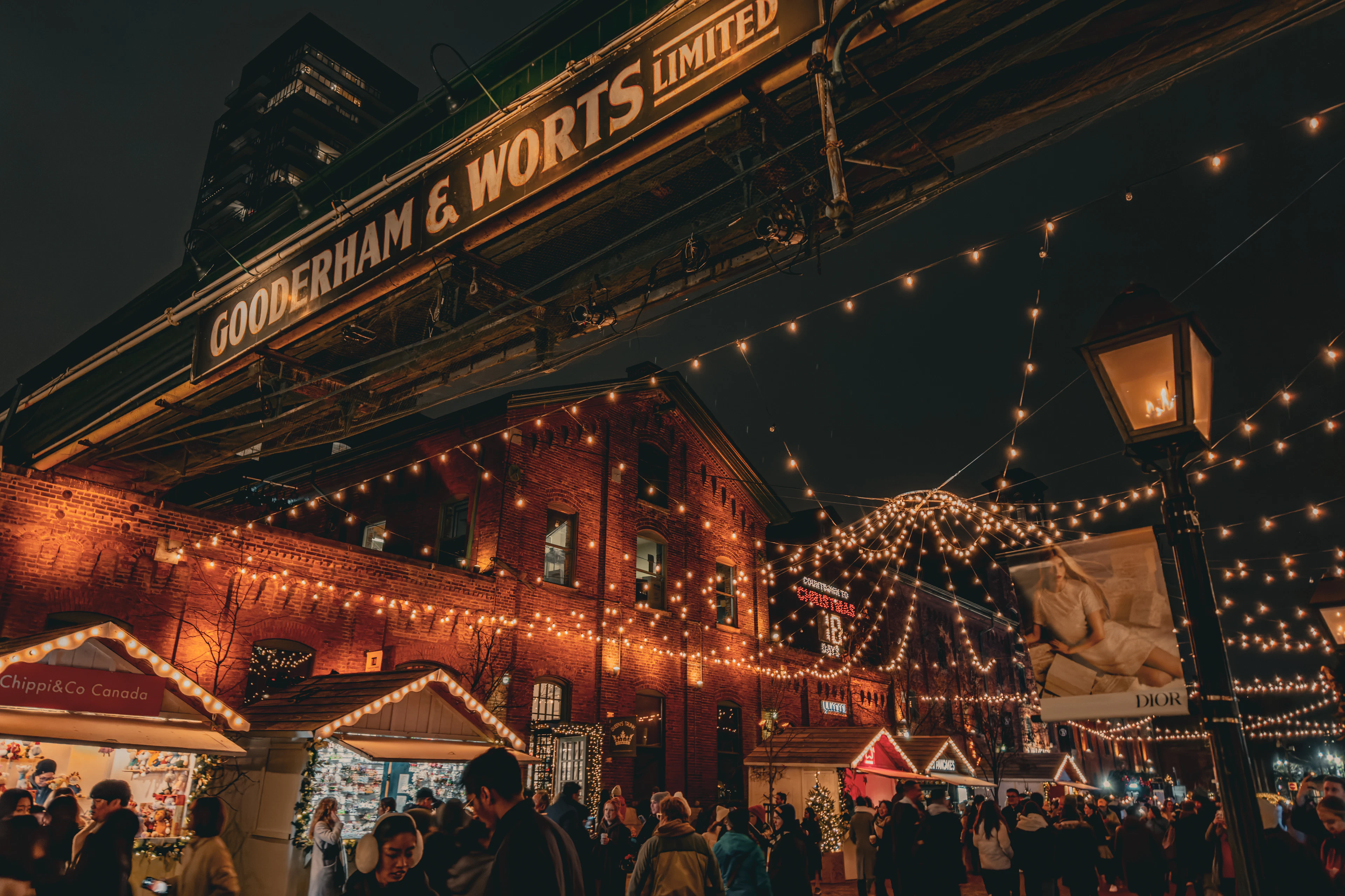 The Distillery District winter market in Toronto at night, with canopies of warm string lights strung between red-brick Victorian warehouses beneath the illuminated Gooderham & Worts sign, and crowds moving among white wooden chalets.