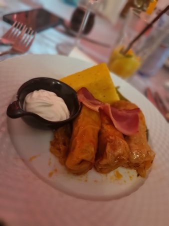 A plate containing three stuffed cabbage rolls with a slice of ham on top, accompanied by a dollop of white sauce in a small black dish. There is a serving of yellow polenta next to the cabbage rolls. The background shows a blurred dining setting with cutlery, a glass, and a drink with a straw.